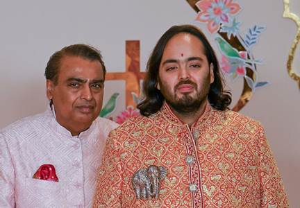 Two Indian men posing for a formal photo: an older man in a white kurta with a red pocket square beside a younger man in a gold, embroidered sherwani, with decorative wall art in the background. - El plan del millonario indio que quería los 80 hipopótamos de Escobar, con los animales silvestres de Venezuela