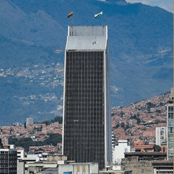 El paisa que terminó detrás de Cien años de soledad y trabajando con J Balvin, Feid y Juanes Tall skyscraper with a dark glass facade and two flags on its roof, set against a mountainous city backdrop. - El paisa que terminó detrás de Cien años de soledad y trabajando con J Balvin, Feid y Juanes