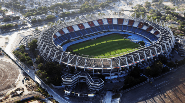 Los constructores detrás de la millonaria remodelación del Estadio Metropolitano de Barranquilla Aerial view of a large oval stadium with blue and red seating and surrounding roads and trees. - Los constructores detrás de la millonaria remodelación del Estadio Metropolitano de Barranquilla