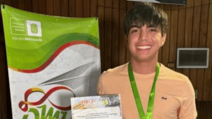 Smiling young man wearing a peach shirt and green medal lanyard holds a certificate at an event, with a colorful banner in the background. - Por segundo año consecutivo, un cucuteño consigue la hazaña de obtener un ICFES perfecto