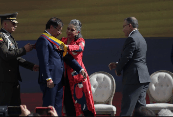 Las jugadas de Iván Name en el Senado que la justicia castigó y pueden perjudicar a María José Pizarro Man in a blue suit receives a yellow sash from a woman in red during a formal award ceremony, with a uniformed officer nearby and a dark backdrop behind them. - Las jugadas de Iván Name en el Senado que la justicia castigó y pueden perjudicar a María José Pizarro