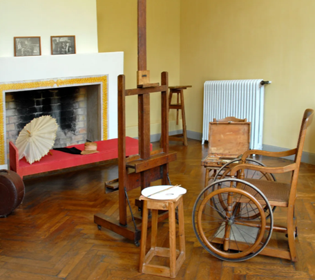 Old art studio with a wooden easel, small stool, and a vintage wheelchair near a yellow wall and brick fireplace. - El día que la esposa de Sarmiento Jr. trajo la unica obra de Renoir que se ha vendido en Colombia
