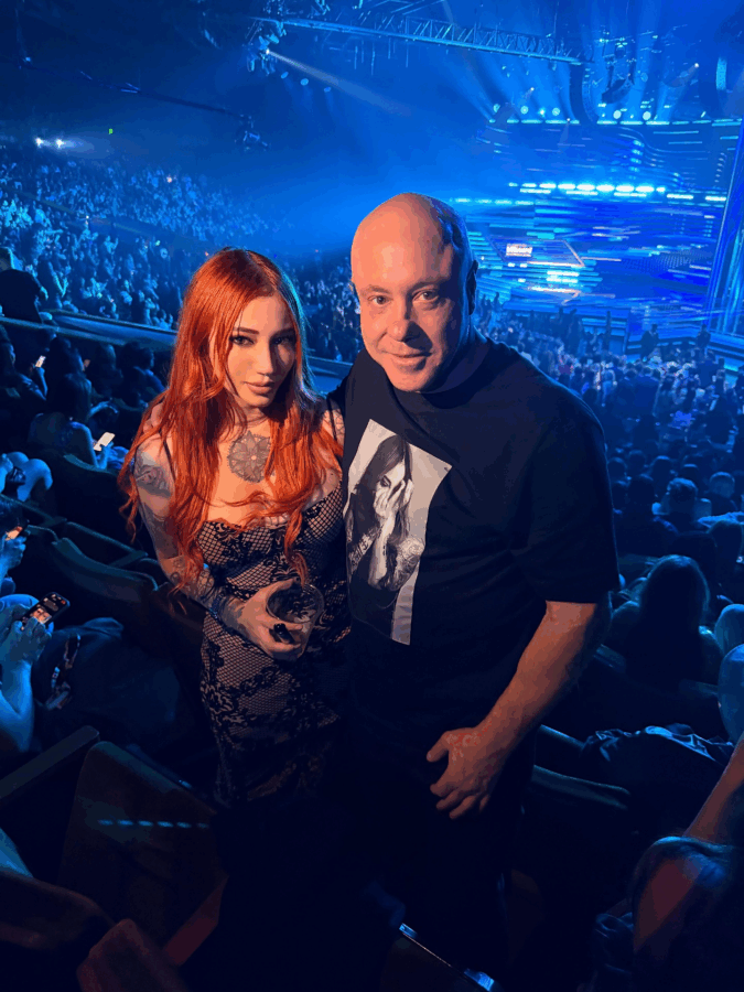Two concertgoers pose for a photo in a blue-lit arena, a red‑haired tattooed woman beside a bald man in a black graphic tee, with a crowded stage behind them. - Quién es Alejandra Rosales, la DJ y periodista que conquistó a Jorge Rausch, famoso jurado de MasterChef