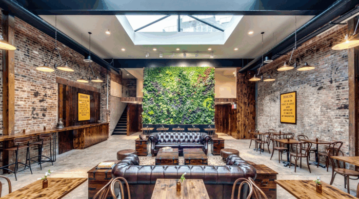 Las cinco empresas cafeteras que apostaron a la estrategia “De la finca a la taza” en 72 horas Industrial cafe interior with brick walls, hanging lights, and a central green living wall flanked by leather sofas and wooden tables. - Las cinco empresas cafeteras que apostaron a la estrategia “De la finca a la taza” en 72 horas