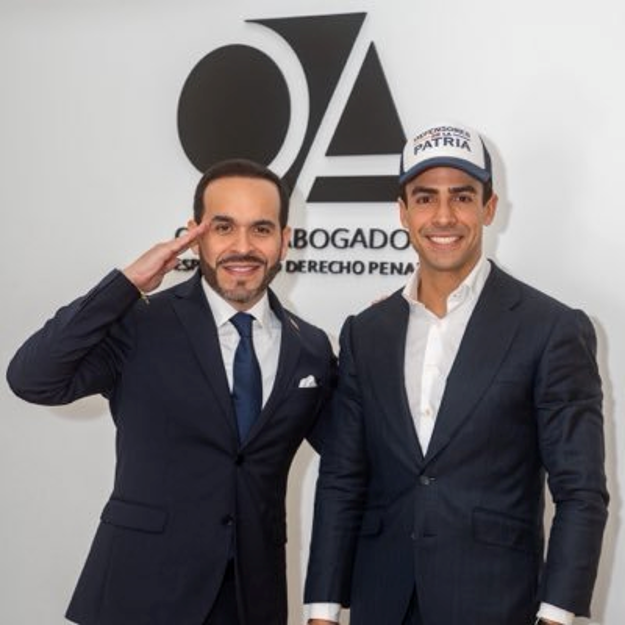 El próximo embajador de Colombia en Bruselas es quien logró cadena perpetua para el asesino del cirujano Edwin Arrieta Two men in navy suits pose and smile in a law office lobby, one giving a salute; behind them is a logo with 'ABOGADOS' and 'DERECHO PENAL' - El próximo embajador de Colombia en Bruselas es quien logró cadena perpetua para el asesino del cirujano Edwin Arrieta