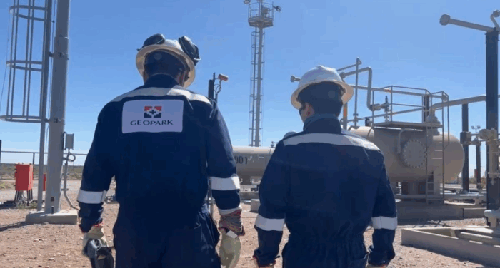 Felipe Bayón, al frente de Geopark le pone el ojo al Magdalena Medio para retomar la senda del fracking Two workers in dark blue coveralls with white stripes stand facing industrial equipment, backs to camera; GEOPARK logo on one jacket under a clear blue sky. - Felipe Bayón, al frente de Geopark le pone el ojo al Magdalena Medio para retomar la senda del fracking