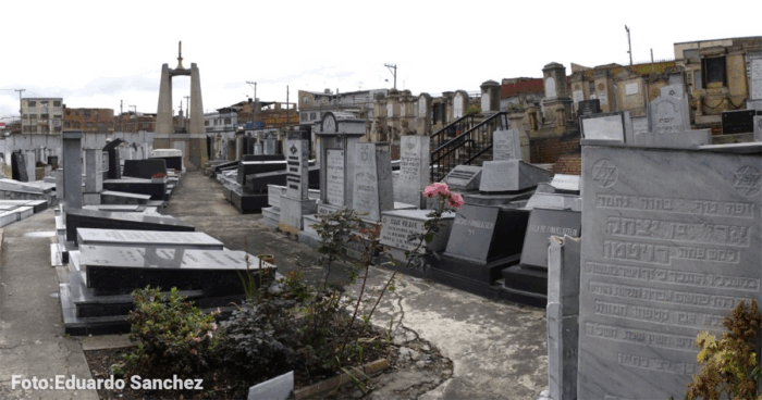 El desconocido cementerio hebreo que guarda 892 tumbas el sur de Bogotá entre talleres de mecánica Wide view of a cemetery with rows of gray tombstones and monuments, a central path, and an urban skyline in the background; pink flowers in the foreground. - El desconocido cementerio hebreo que guarda 892 tumbas el sur de Bogotá entre talleres de mecánica