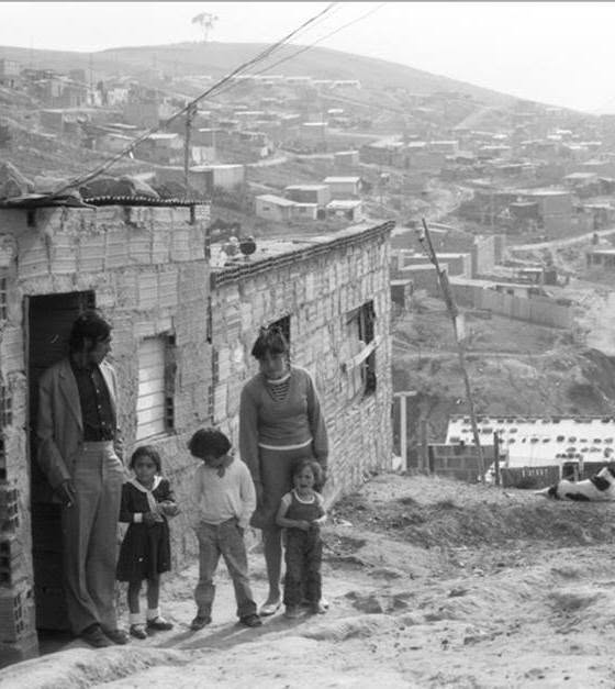 Family of five stands outside a brick house on a dirt path in a hillside shantytown; dense housing rises in the background. - Esta es la historia de Potosí, el barrio de Ciudad Bolívar donde muchos le tienen miedo a caminar sus calles