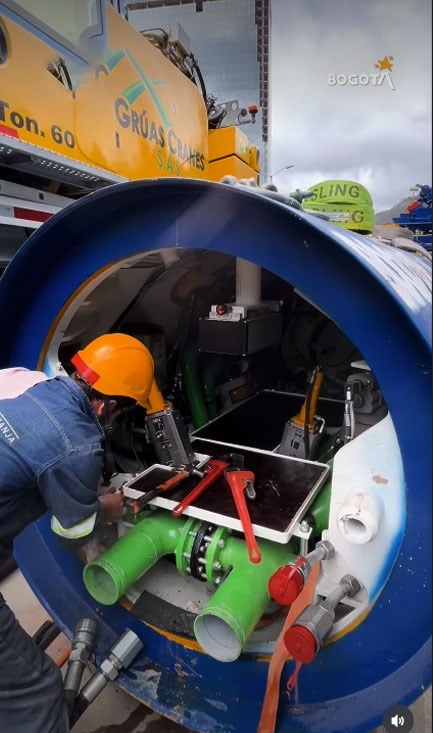 Quién es Carmen, la tuneladora alemana que reduce el trabajo de 2 años a tan solo 10 meses en la carrera séptima Worker in an orange hard hat and safety vest assembles or inspects green pipes inside a large blue cylindrical tank, with red-handled tools nearby. - Quién es Carmen, la tuneladora alemana que reduce el trabajo de 2 años a tan solo 10 meses en la carrera séptima