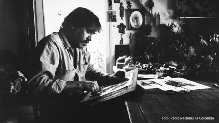 Cuando París, hace 50 años, fue el semillero de 29 grandes artistas colombianos Man seated at a cluttered desk sketches in a large book, in a dimly lit room with papers and plants; photo credit reads 'Foto: Radio Nacional de Colombia' - Cuando París, hace 50 años, fue el semillero de 29 grandes artistas colombianos