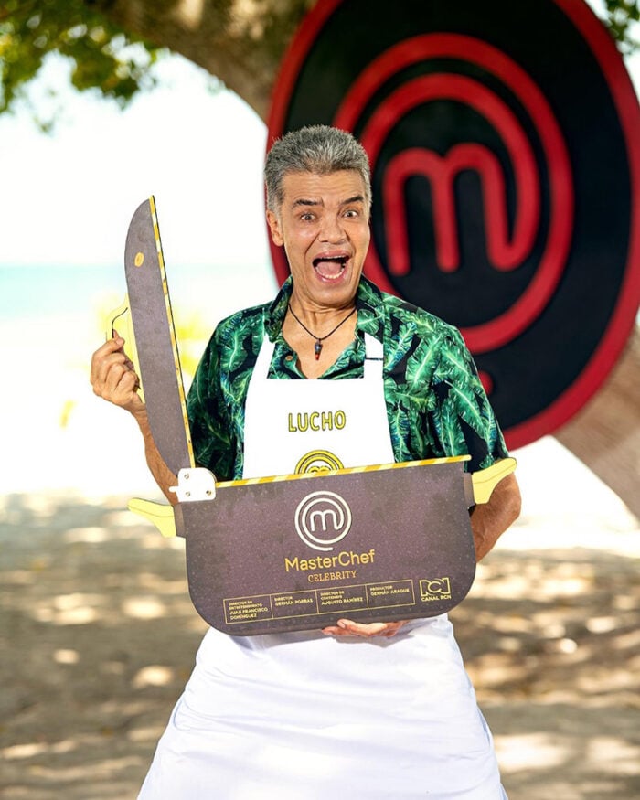 Luis Eduardo Arango, el actor paisa que se resiste a dejar la actuación en la que lleva más de medio siglo Celebrity MasterChef contestant Lucho on a beach, cheerfully holding a large MasterChef prize box with a big logo in the background. - Luis Eduardo Arango, el actor paisa que se resiste a dejar la actuación en la que lleva más de medio siglo