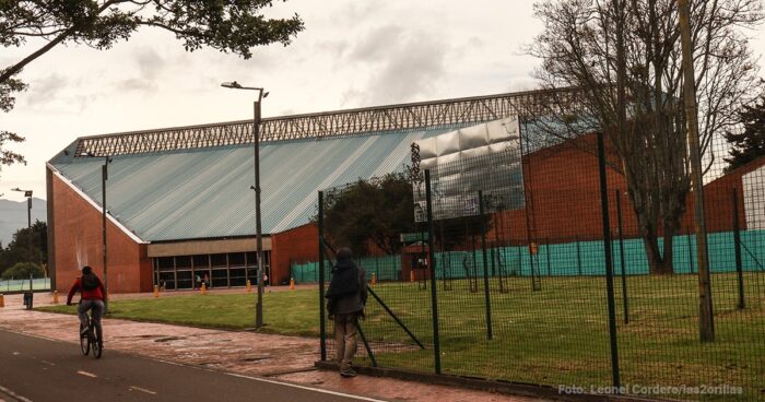 Después de diecisiete años y nueve contratos por más de .000 millones la Unidad Deportiva Salitre sigue igual Cyclist riding along a path beside a brick building with a slanted light-blue roof and a tall fence. - Después de diecisiete años y nueve contratos por más de .000 millones la Unidad Deportiva Salitre sigue igual