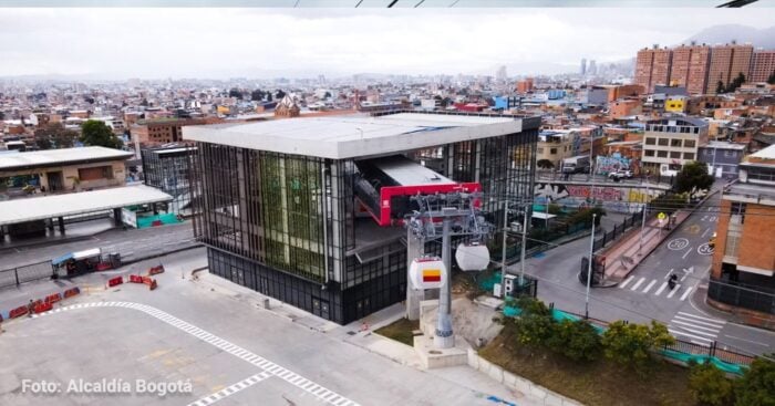 Dos familias ligadas al transporte bogotano junto a los bolivianos de Mi Teleférico operarán el cable de San Cristóbal Aerial view of a modern cable car station in Bogotá with a red gondola, set among dense urban buildings and hills in the distance. - Dos familias ligadas al transporte bogotano junto a los bolivianos de Mi Teleférico operarán el cable de San Cristóbal