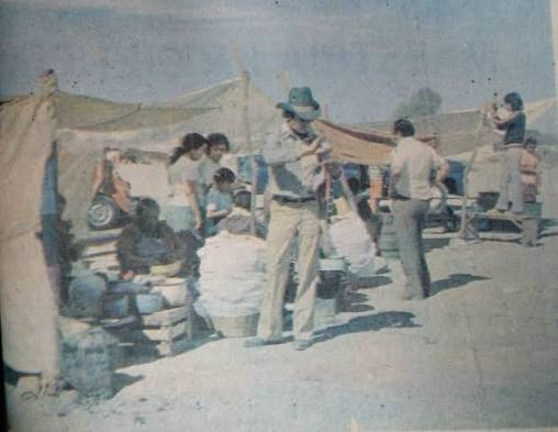 Desert camp scene with canvas tents, people gathered around crates and talking near a dusty settlement. - Esta es la historia de Potosí, el barrio de Ciudad Bolívar donde muchos le tienen miedo a caminar sus calles