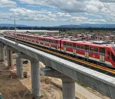 Red passenger train on an elevated track passing over a construction area, with a town and hills in the distance under a partly cloudy sky. - Que hicieron y que dejaron de hacer los 3 presidentes y 4 últimos Alcaldes de Bogotá por el Metro