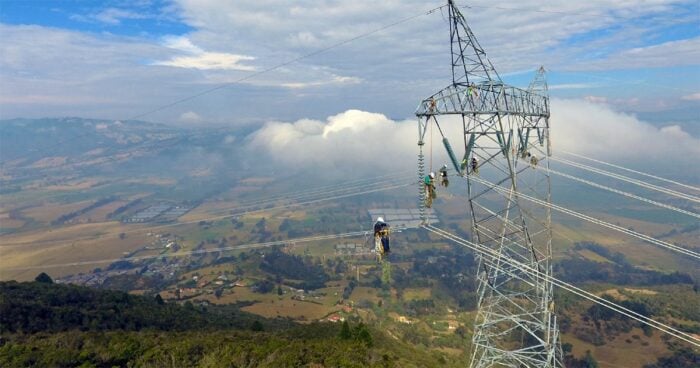 Después de 10 años se logrará al fin enviar energía a Bogota a través de un conector que une a Boyacá y Santander Workers in safety gear climbing a tall transmission tower to inspect power lines over a valley. - Después de 10 años se logrará al fin enviar energía a Bogota a través de un conector que une a Boyacá y Santander