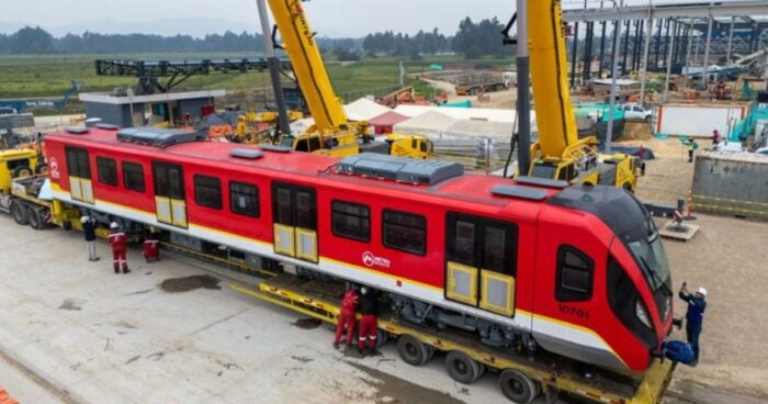 Red passenger train car on a specialized trailer, cranes lifting it at a construction site with workers nearby on a paved area. - Que hicieron y que dejaron de hacer los 3 presidentes y 4 últimos Alcaldes de Bogotá por el Metro