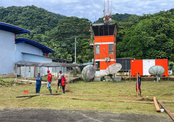 Luego de 4 años de retrasos, adiciones y mil millones invertidos Bahía Solano, en Chocó, estrena aeropuerto Group of workers in blue and red coveralls raking and shoveling on a grassy field near an orange control tower and several large satellite dishes.