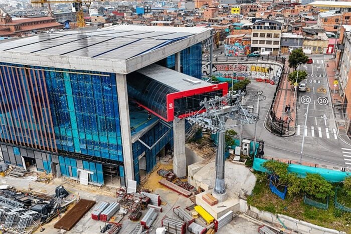 Dos familias ligadas al transporte bogotano junto a los bolivianos de Mi Teleférico operarán el cable de San Cristóbal Aerial view of a glass-front building under construction with a red cable-car station and elevated rails across a city plaza. - Dos familias ligadas al transporte bogotano junto a los bolivianos de Mi Teleférico operarán el cable de San Cristóbal