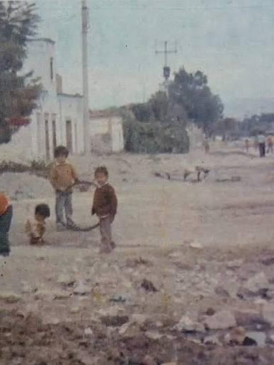 Three children stand on a rocky dirt street in a residential area, with low white buildings and trees in the background. - Esta es la historia de Potosí, el barrio de Ciudad Bolívar donde muchos le tienen miedo a caminar sus calles