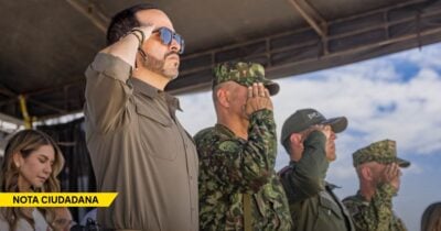 Group of uniformed men saluting on an outdoor stage; a yellow NOTA CIUDADANA banner is visible in the corner. - Las2orillas.co: Historias, voces y noticias de Colombia