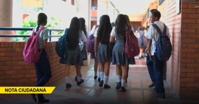 Group of students in school uniforms walking down a brick hallway with backpacks; yellow 'NOTA CIUDADANA' banner bottom-left. - Las2orillas.co: Historias, voces y noticias de Colombia