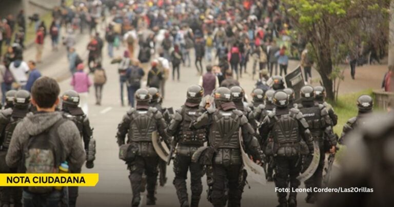 Riot police in full armor march toward a crowded street filled with protesters; a man with a backpack stands in the foreground. - Ni represión a los marchantes ni impunidad a los vándalos: así debe regularse la protesta