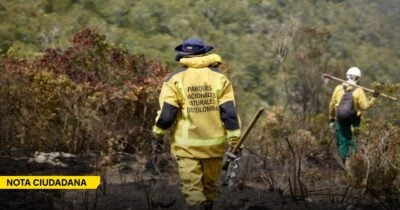 Two firefighters in yellow protective gear walk through a burned, smoky landscape, carrying tools after a wildfire. - Las2orillas.co: Historias, voces y noticias de Colombia