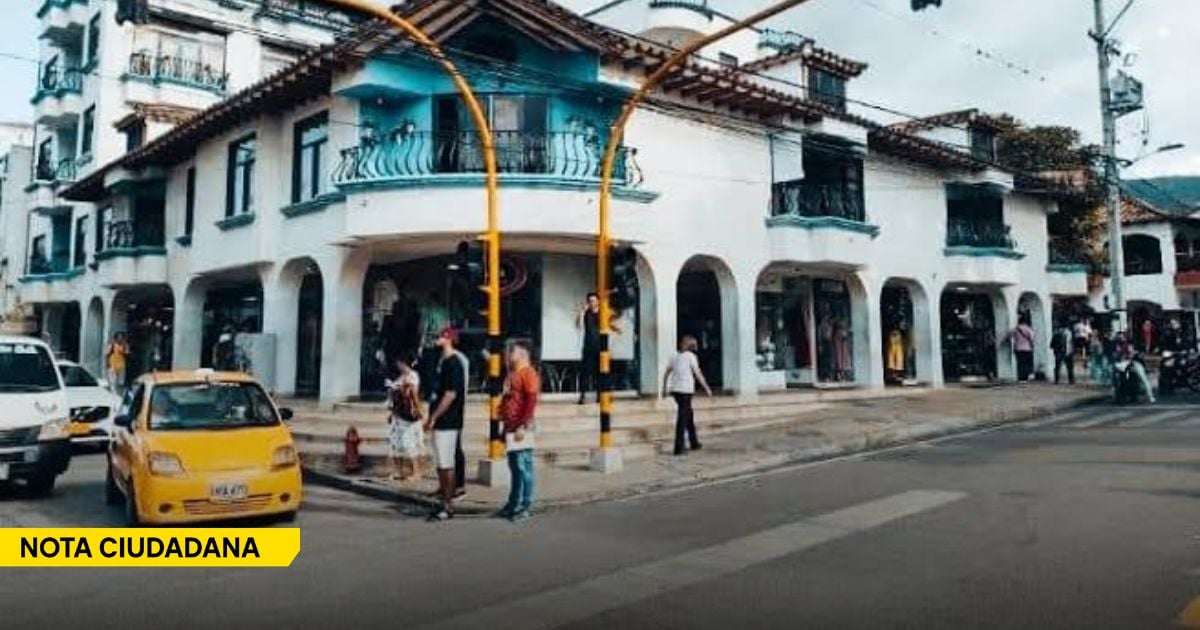 Street corner scene with a white arched building, pedestrians on the sidewalk, and a yellow taxi in the foreground; a yellow banner reads NOTA CIUDADANA off to the side. - Entre habitantes de calle misteriosamente desaparecidos y campesinos extorsionados, el miedo se apodera de Ocaña