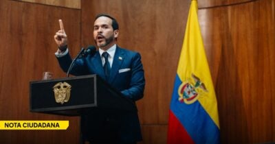 Man in a navy suit speaks at a podium, raising his right index finger, with a microphone in front and a Venezuelan flag in the background. - Las2orillas.co: Historias, voces y noticias de Colombia