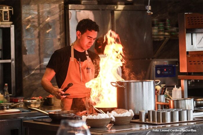 El barrio más ‘cool’ de América Latina lo tiene Bogotá y vivir allá no es barato Chef in a busy kitchen standing at a stove with a large flare from a pan, wearing an apron and black shirt, using tongs. - El barrio más ‘cool’ de América Latina lo tiene Bogotá y vivir allá no es barato