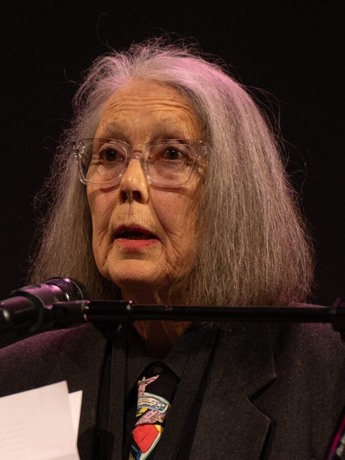 Anne Carson, la poeta que apagó el celular cuando le dieron el Nobel de Literatura y donará .800 millones del premio Older woman with long gray hair and glasses speaking into a microphone on stage.