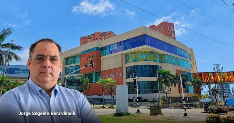 Las2orillas.co: Historias, voces y noticias de Colombia A man in a light blue button-up shirt stands in the foreground with a colorful modern shopping mall in the background. - Las2orillas.co: Historias, voces y noticias de Colombia