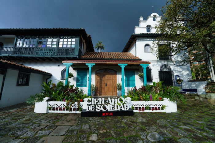 Así fue como Netflix convirtió a Macondo en un negocio que le dejó más de medio billón de pesos a Colombia White colonial house with turquoise doors and a garden, a large 'Cien Años de Soledad' sign in front and Netflix logo.