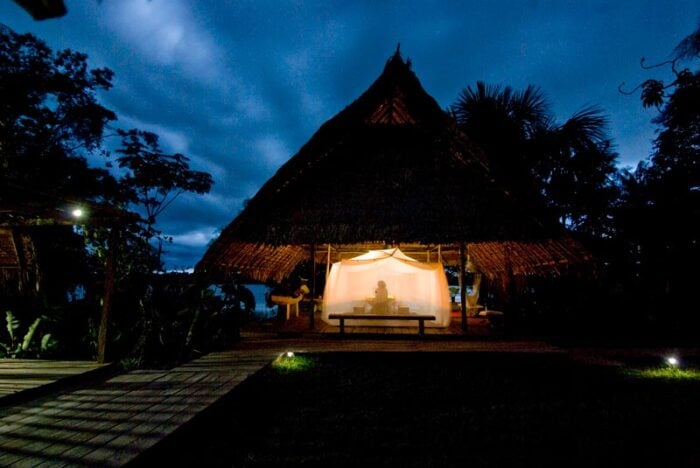 El hotel de lujo que el pintor Diego Samper levantó en la selva del Amazonas desde hace 40 años Thatched open-air hut on a beach at night with warm light inside and a person seated under a sheer curtain canopy - El hotel de lujo que el pintor Diego Samper levantó en la selva del Amazonas desde hace 40 años