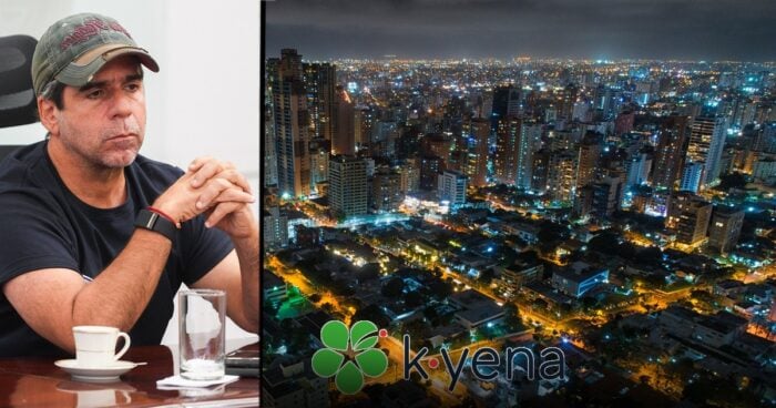 Man in a cap sits at a table, with a nighttime cityscape and a kyena logo on a large screen beside him. - Las2orillas.co: Historias, voces y noticias de Colombia