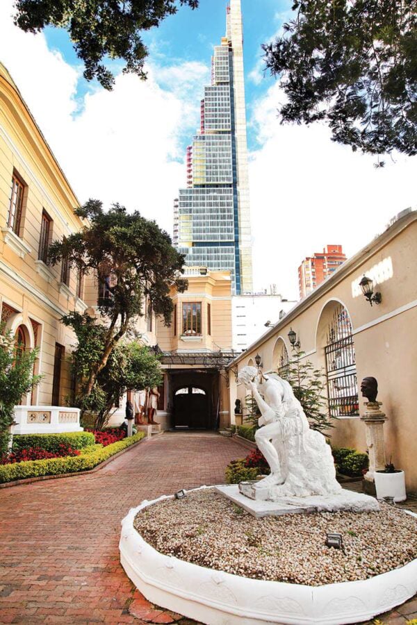 Courtyard with manicured garden, a white statue fountain, and a tall modern skyscraper under construction in the background. - Así es la mansión en Bogotá de Leo Kopp, el fundador de Bavaria, que terminó convertida en el templo de los masones
