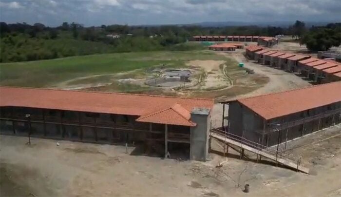 La Federación de Cafeteros se mete al negocio hotelero con un megaproyecto frente al Parque del Café Aerial view of a large compound with long red-tiled buildings forming a U-shaped perimeter around a central circular yard, with dirt ground and surrounding greenery under a cloudy sky. - La Federación de Cafeteros se mete al negocio hotelero con un megaproyecto frente al Parque del Café