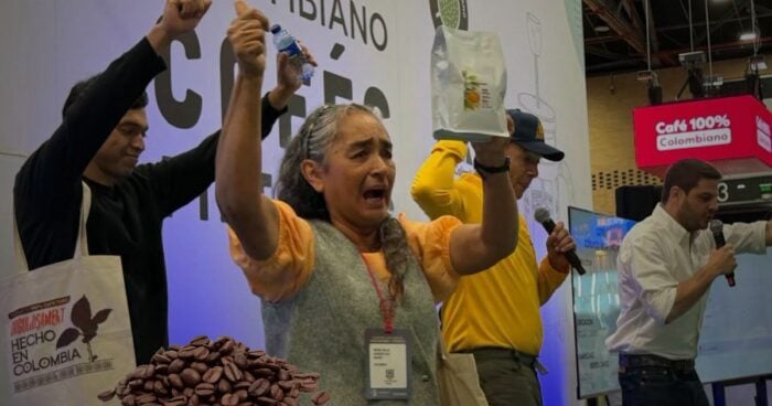Group of people on a stage at a coffee event, raising their hands in celebration with a bag and bottle, beans visible in the foreground. - Una caficultora del Cauca que lo perdió todo en un incendio vendió su última cosecha a 400 dólares por libra