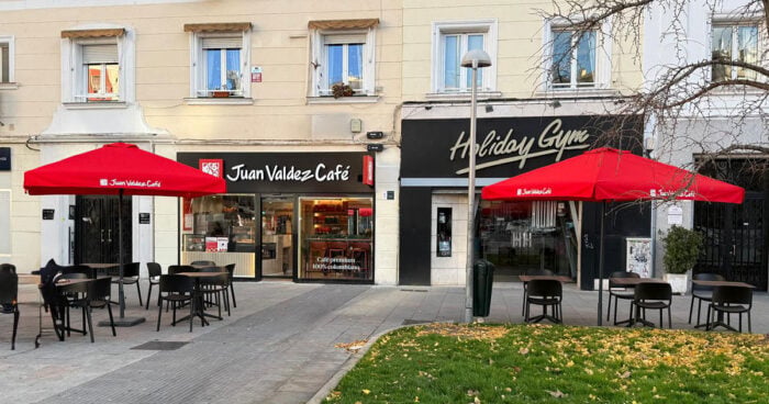 La jugada de Omar González con el grupo Trinity para llevar las tiendas Juan Valdez a Cataluña: abrirá diez en Barcelona Outdoor cafe seating with red umbrellas in front of Juan Valdez Café and a storefront labeled Holiday Gym on a sunny street.