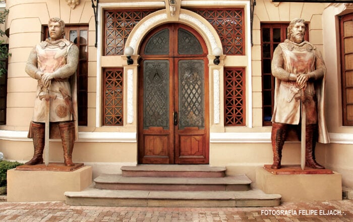 Two life-size knight statues flank a carved wooden arched door on a beige building, swords sheathed, with steps leading up to the entrance. - Así es la mansión en Bogotá de Leo Kopp, el fundador de Bavaria, que terminó convertida en el templo de los masones