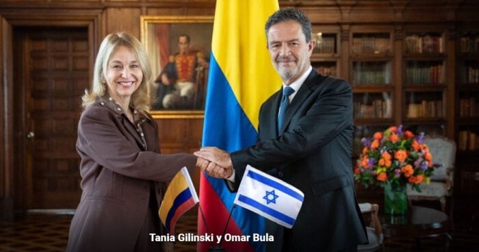Tania Gilinski and Omar Bula shake hands in a formal room, with the Colombian and Israeli flags behind them and a flower arrangement nearby. - Las2orillas.co: Historias, voces y noticias de Colombia