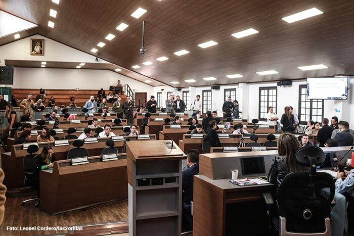 Indignación por el polémico contrato que firmó el Concejo de Bogotá para lavarle los carros a los concejales A crowded council chamber or lecture hall with attendees seated at wooden desks facing a central podium. - Indignación por el polémico contrato que firmó el Concejo de Bogotá para lavarle los carros a los concejales