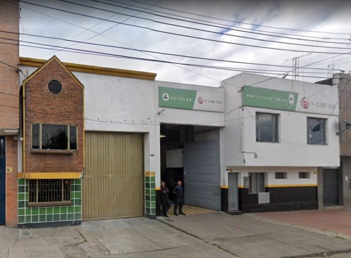 Indignación por el polémico contrato que firmó el Concejo de Bogotá para lavarle los carros a los concejales Facade of an auto repair workshop with an open bay door, signage reading AUTOLAB and V-CARS S.A.S., and two workers standing at the entrance. - Indignación por el polémico contrato que firmó el Concejo de Bogotá para lavarle los carros a los concejales