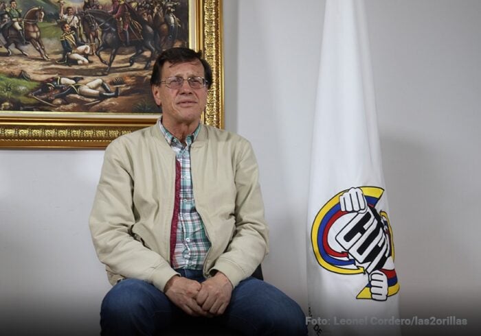 Por qué pelean entre ellos los 21 miembros del comité ejecutivo nacional de la Central Unitaria de Trabajadores Man seated in a beige jacket and plaid shirt, wearing glasses, with a flag bearing a colorful circular logo beside him. - Por qué pelean entre ellos los 21 miembros del comité ejecutivo nacional de la Central Unitaria de Trabajadores