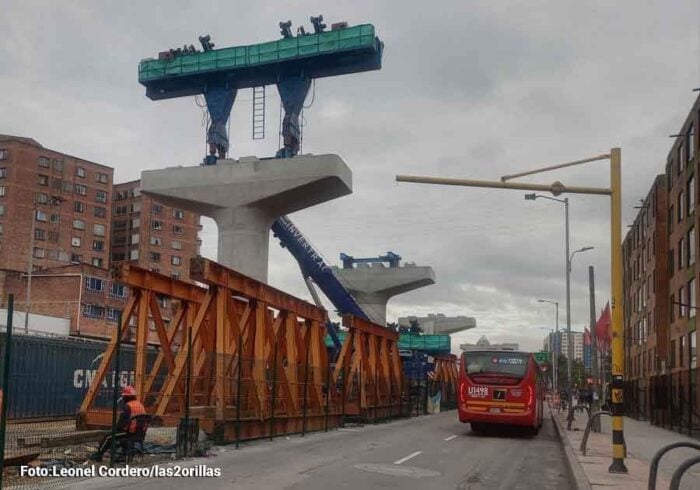 Luz verde del Tigre para que los bogotanos empiecen a soñar con la Línea 4 del metro Urban construction zone on a city street building an elevated structure: concrete supports with green segments, orange steel frames, and a red bus passing by. - Luz verde del Tigre para que los bogotanos empiecen a soñar con la Línea 4 del metro