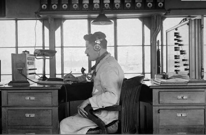 La vieja estación del tren levantada hace 140 años que aún sobrevive en el corazón de Fontibón Vintage telephone switchboard operator seated at a desk with an old headset, in a room filled with early communications equipment and a large window behind. - La vieja estación del tren levantada hace 140 años que aún sobrevive en el corazón de Fontibón