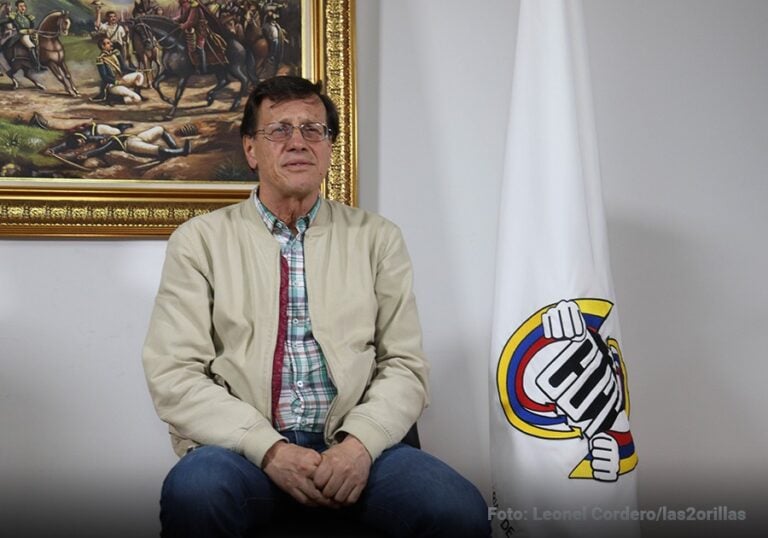 Las2orillas.co: Historias, voces y noticias de Colombia Man seated in a casual beige jacket and plaid shirt, facing the camera, with a flag featuring a clenched-fist logo to his right and a framed painting behind him. - Las2orillas.co: Historias, voces y noticias de Colombia