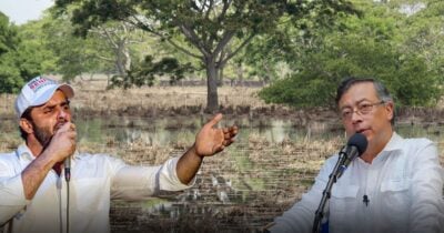 Las2orillas.co: Historias, voces y noticias de Colombia Two men in white shirts speak into microphones outdoors, one gesturing with an outstretched arm as they stand by a pond and trees. - Las2orillas.co: Historias, voces y noticias de Colombia