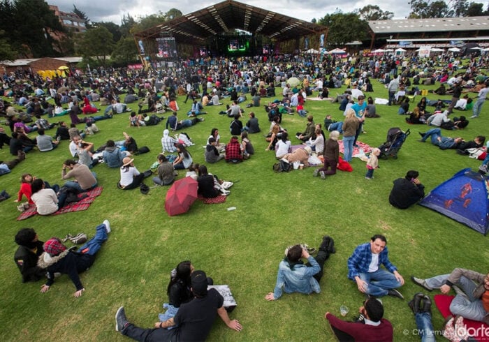 Mientras Rock al Parque celebra 30 años, la entidad que lo maneja lleva una década bajo la lupa de la Contraloría Crowd lounging on grass at an outdoor festival with a large stage and screens in the background. - Mientras Rock al Parque celebra 30 años, la entidad que lo maneja lleva una década bajo la lupa de la Contraloría